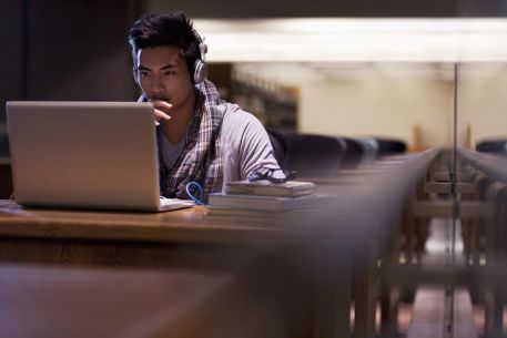 A student sitting in a library with headphones on while working on a laptop.