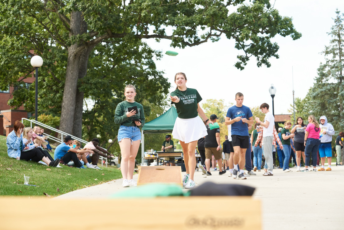 Two girls smiling while playing cornhole on a sidewalk with trees and a crowd of people in the background