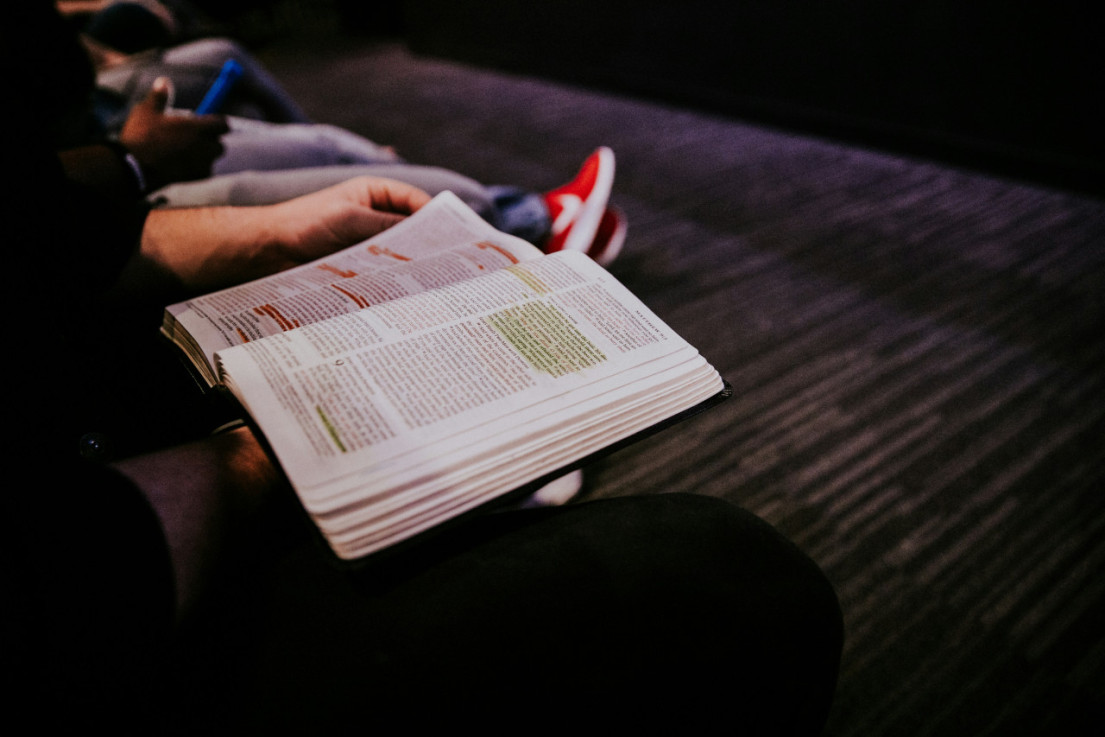 A church attender holds a Bible open to a page in the Gospel of Matthew. The legs of other people sitting in the same row of chairs in the church auditorium are visible.