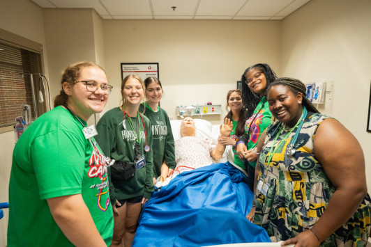 Six summer nursing students pose with a medical dummy in a hospital bed.