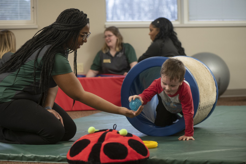 An HU OTD Student works with a small child in a therapy environment. There are other students in the background.