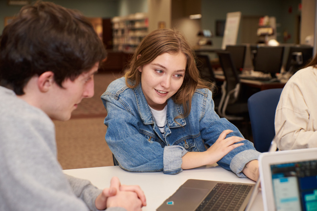 Two students, one male and one female, look together at a laptop. They are sitting at a table on the main level of RichLyn Library on the campus of Huntington University. 