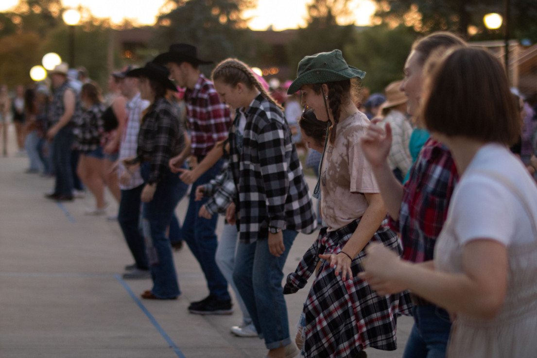 Male and female Huntington University students stand in a line during the Hoedown on the campus of Huntington University. They are dancing as the sun sets. Most of them are looking at their feet to make sure they are getting the steps correct.