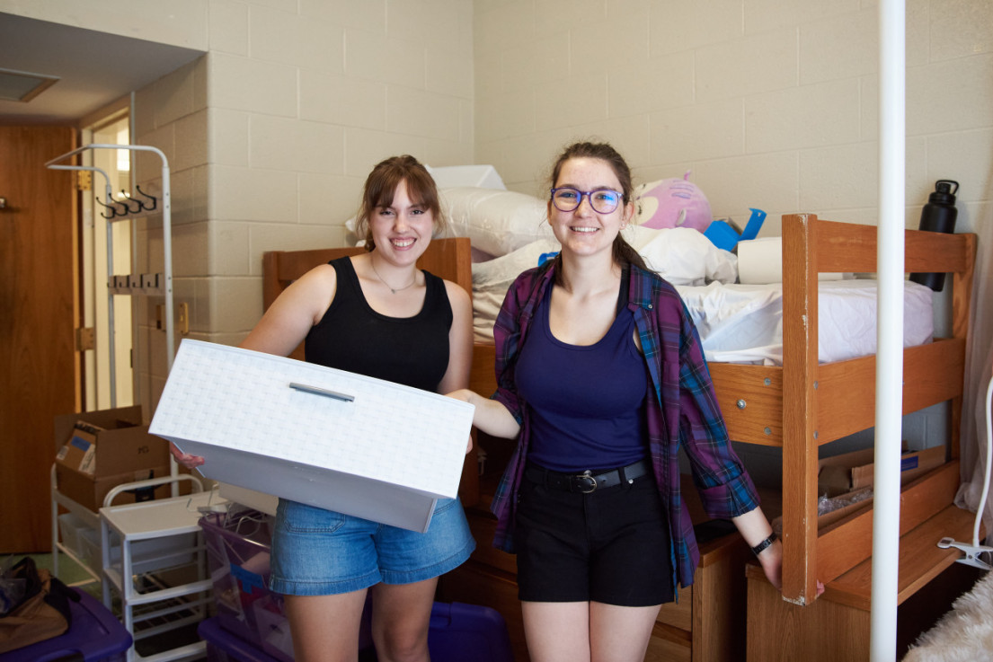 Two female students standing in a residence hall room during move-in surrounded by containers and other belongings